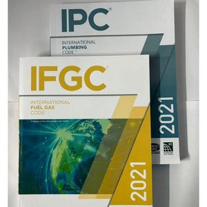 Op de afbeelding: Twee boeken, getiteld "IPC INTERNATIONAL PLUMBING CODE" en "IFGC INTERNATIONAL FUEL GAS CODE", zijn op elkaar gestapeld. Het IFGC-boek heeft een wereldkaartafbeelding en de tekst "2021". Het logo van de American Gas Association is zichtbaar.