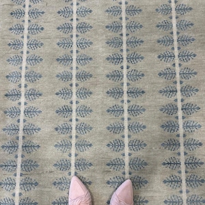 May include: A beige rug featuring a repeating pattern of blue leaf-like designs and white vertical stripes. The rug has a textured look. Pink slip-on shoes are visible at the bottom of the image.