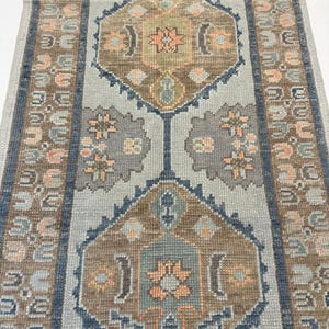 May include: A rectangular rug with a central design featuring geometric shapes in shades of brown, peach, and blue. The rug has a light blue background with a border of repeating patterns in brown and blue. The rug is suitable for a hallway or living room.