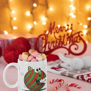 May include: A white mug filled with marshmallows, featuring a cartoon Christmas ornament design with red and green stripes. Candy canes and a red "Merry Christmas" sign are also visible, creating a festive holiday scene.