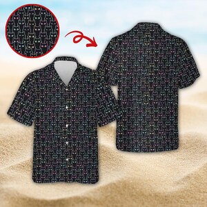 May include: Black button-down shirt with a repeating cat face pattern. The cat faces have colorful outlines and eyes. The shirt has short sleeves and a collared neckline. The background is a sandy beach with a blue sky.