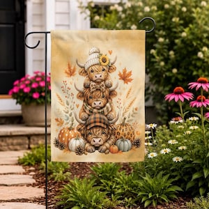 May include: A decorative garden flag featuring a vertical stack of three fluffy Highland cows in a warm, autumnal colour palette. The cows are adorned with a knit hat, a sunflower, a bow tie, a scarf, and a plaid cap, surrounded by pumpkins and leaves.