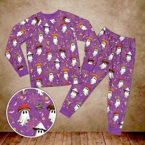 Könnte beinhalten: Ein langärmeliges Schlafanzug-Set aus Oberteil und Hose in Lila mit einem durchgehenden Muster aus weißen Geistern, die verschiedene bunte Pilze als Hüte tragen, durchsetzt mit orangefarbenen Spinnenweben und kleinen Fledermäusen auf dem Stoff.