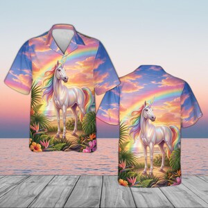 May include: A short-sleeved button-down shirt featuring a vibrant illustration of a white unicorn with a rainbow mane and tail, set against a sunset backdrop with a rainbow. Tropical flowers and foliage frame the scene.