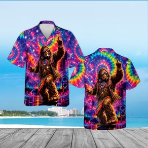 May include: A short-sleeve button-down shirt featuring a vibrant tie-dye pattern in shades of purple, blue, pink, and yellow. The graphic print depicts a brown, furry creature wearing sunglasses and headphones, set against a colorful background.