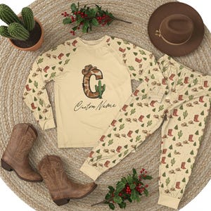 May include: A beige pajama set with a cowboy theme, featuring a long-sleeved top and pants. The top has a large letter "C" with a cowboy hat and cactus design, with "Custom Name" below. The pants and sleeves have a repeating pattern of cowboy boots, hats, and cacti. A brown cowboy hat and boots are also present.