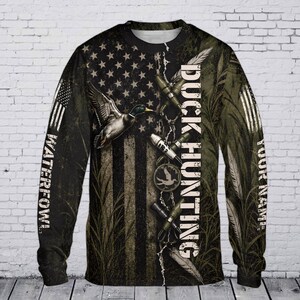May include: A long-sleeve shirt featuring a dark, distressed American flag pattern with a flying mallard duck, shotgun shells, and tall grass graphics. The text DUCK HUNTING is printed vertically, with WATERFOWL and YOUR NAME on the sleeves.