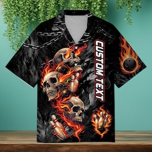 May include: Black bowling shirt with a skull and flame design. The shirt features a v-neck collar and short sleeves. The graphic includes skulls, bowling pins, and bowling balls engulfed in flames, with the text "CUSTOM TEXT" on the side.