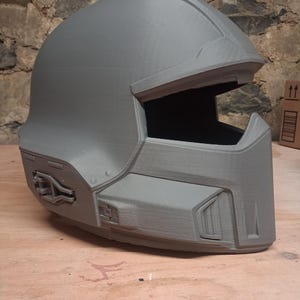 Könnte beinhalten: Ein grauer, 3D-gedruckter Integralhelm mit einer glatten, abgerundeten Oberseite, einer breiten horizontalen Visieröffnung und kantigen geometrischen Details an den Seiten und im Kinnbereich. Die Oberfläche zeigt feine horizontale Schichtlinien.