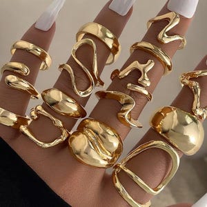 Puede incluir: Un conjunto de anillos dorados con diseños variados, incluyendo bandas lisas y formas abstractas. Los anillos se exhiben en dedos con uñas largas pintadas de blanco, creando una estética elegante y a la moda.