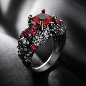 May include: A gothic-style ring with a black band, featuring a large, square-cut red gemstone and two smaller oval red stones. The ring is adorned with skulls, leaf-like details, and small, sparkling white stones.
