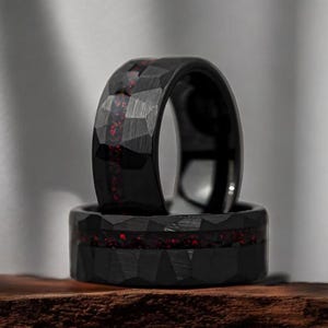Puede incluir: Dos anillos negros con superficie facetada. Los anillos tienen una incrustación roja moteada. Los anillos están apilados uno encima del otro, con el anillo superior ligeramente inclinado. Los anillos se exhiben sobre una superficie de madera.