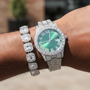Op de afbeelding: Een zilverkleurig horloge met een groene wijzerplaat en een bijpassende armband, beide bezet met heldere edelstenen. Het horloge heeft Romeinse cijfers en een datumweergave. De armband heeft vierkante stenen.