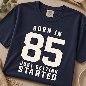May include: A navy blue short-sleeve t-shirt featuring the white text BORN IN 85 JUST GETTING STARTED in a bold, athletic font. The shirt has a crew neckline and a Comfort Colors brand label printed on the inside of the collar area.