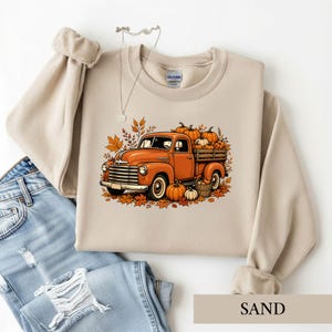 May include: A sand-colored crewneck sweatshirt featuring a graphic print of an orange vintage pickup truck loaded with pumpkins and surrounded by autumn leaves. The Gildan brand tag is visible at the inner neckline, and a silver necklace is present.