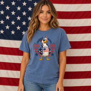 Woman wearing America 250th anniversary shirt featuring patriotic eagle design and 1776 2026 graphic in front of American flag backdrop.
