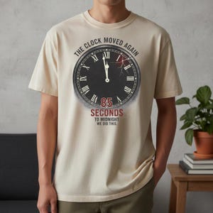May include: Beige t-shirt with a distressed clock graphic. The clock face shows Roman numerals and the text "THE CLOCK MOVED AGAIN." Below the clock, it reads "85 SECONDS TO MIDNIGHT WE DID THIS."