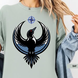 May include: A light green t-shirt featuring a stylized black bird with outstretched wings, a red eye, and a blue and white circular design. Above the bird is a blue and white compass rose. The word "BAY" is visible on the bottom right.