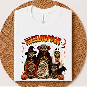 May include: A white t-shirt featuring a graphic of six snakes dressed in Halloween costumes, including a witch, vampire, ghost, pumpkin, mummy, and bat. The text HISSINGPOOK is printed in orange and yellow above the snakes, with small bats and stars.