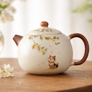 Puede incluir: Tetera de cerámica color crema con asa, boquilla y pomo de tapa de madera. La tetera presenta un diseño pintado a mano de un gato, flores amarillas y hojas verdes. La tetera está sobre una mesa de madera.