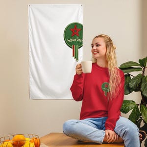 Puede incluir: Una bandera blanca con un emblema verde y rojo con una estrella y texto árabe. Se ve una sudadera roja con un emblema a juego. La bandera y la sudadera comparten un diseño similar, lo que sugiere un tema de orgullo nacional o identidad cultural.