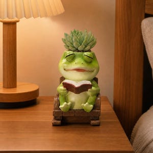 Puede incluir: Una figura de rana verde sentada en una mecedora marrón mientras sostiene un libro abierto. La rana tiene una planta suculenta de color verde claro que crece en la parte superior de su cabeza, con los ojos cerrados y una leve sonrisa.