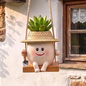 May include: A hanging planter shaped like a smiling character wearing a straw hat, holding a small metal shovel, and sitting on a wooden swing. The planter contains a green succulent plant and is suspended by two ropes from a wooden structure.