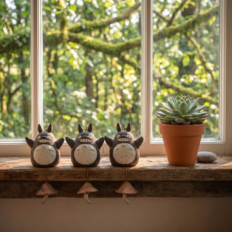 Puede incluir: Tres figuras de cer&aacute;mica gris oscuro con vientres blancos y orejas puntiagudas, cada una con un dije en forma de seta colgando de la base. Junto a las figuras hay una planta suculenta verde en una maceta de terracota y una peque&ntilde;a piedra gris.