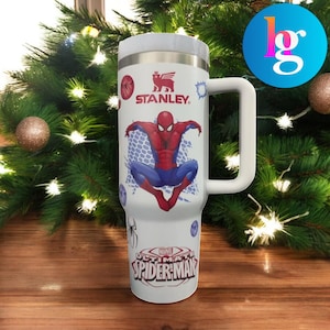 A stainless steel tumbler mug featuring a Spider-Man design, placed on a wooden surface with a Christmas tree in the background.