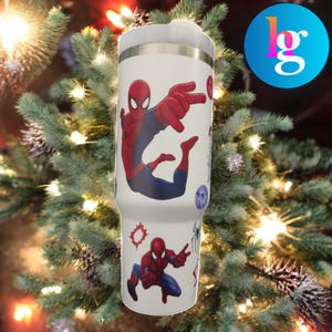 A Spiderman-themed tumbler or thermos mug is displayed against a backdrop of a Christmas tree with twinkling lights.