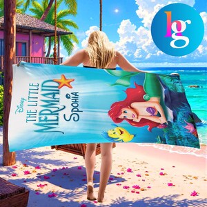 A woman is walking on a beach holding a large towel featuring the character The Little Mermaid from the Disney film.