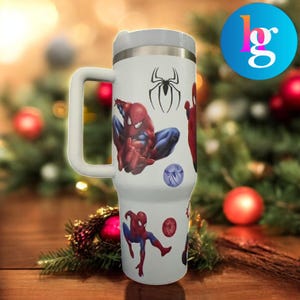 A white travel mug with a Spiderman design printed on it, placed on a wooden surface with Christmas decorations in the background.