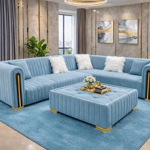 以下が含まれることがあります： ライトブルーのコーナーソファと、お揃いのオットマンとラグ。 ソファはリブのデザイン、ゴールドのアクセント、白のふわふわクッションが特徴です。 オットマンは房状のトップとゴールドの足が付いています。 部屋にはゴールドのトリムとアートワークがあります。