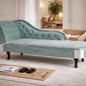Può includere: Una chaise longue turchese con bottoni e gambe in legno scuro. Il mobile è avvolto in plastica protettiva. Uno scatolone e un tappeto sono sullo sfondo. Un tavolino con accessori è in primo piano.