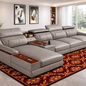 Puede incluir: Un gran sofá seccional moderno en color gris neutro. El sofá cuenta con mesas laterales integradas con puertos de carga y portavasos. Se encuentra sobre una alfombra roja estampada. El sofá tiene una chaise longue y un respaldo acolchado.