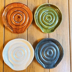 Könnte beinhalten: Vier Keramik-Löffelablagen in Orange, Grün, Weiß und Dunkelblau. Jede Ablage hat ein Spiralmuster in der Mitte und einen gebogenen Rand. Sie werden auf einer Holzoberfläche präsentiert. Maße in Zentimetern.