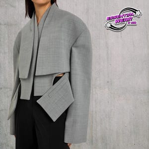 May include: A light gray cropped jacket with a unique design, featuring a draped collar and a rectangular pocket. The jacket is part of the "Essential Wear" collection, as indicated by the logo in the upper right corner. The model is wearing black pants.
