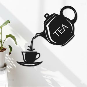 May include: Black metal wall art depicting a teapot pouring tea into a teacup on a saucer. The word "TEA" is displayed on the teapot. The art is mounted on a white wall, with a potted plant visible in the background.