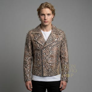 May include: A brown and tan snakeskin-patterned leather jacket with silver metal spikes on the shoulders and collar. The jacket has a classic biker style with a zipper closure and multiple pockets. The model is wearing a white t-shirt and black pants.