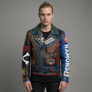 Pode incluir: Jaqueta de couro preta com detalhes azuis e vermelhos, adornada com tachas douradas e pontas prateadas. A manga exibe "RAMONS", enquanto os patches dizem "FUCK WAR" e "DISCHARGE". Uma peça de moda ousada e rebelde.