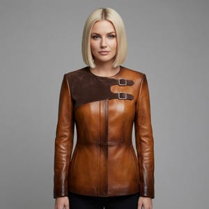 May include: A brown leather jacket with a contrasting dark brown suede shoulder panel. The jacket features two silver buckles and a tailored fit. The leather has a gradient effect, transitioning from lighter to darker shades of brown.