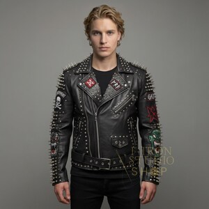 May include: Black leather jacket with silver studs and spikes. The jacket features multiple patches with skull, cross, and other graphic designs. It has a zipper closure and a belt at the waist. The model is wearing a black t-shirt and black pants.
