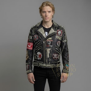 Könnte beinhalten: Schwarze Lederjacke mit goldfarbenen Stacheln und Aufnähern. Die Jacke hat einen Reißverschluss und mehrere Aufnäher mit Totenkopf- und anderen grafischen Designs. Der Text "F CKI THE CISTEM" ist sichtbar. Getragen über einem schwarzen T-Shirt.