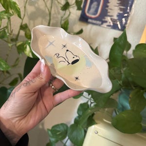 Puede incluir: Un plato de cerámica color crema con bordes ondulados con un diseño de Snoopy. Snoopy está representado durmiendo sobre una luna creciente amarilla, con nubes azules y estrellas. El plato se sostiene en una mano, rodeado de vegetación.
