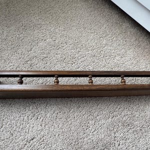 May include: A dark brown wooden shelf with a raised back and decorative spindles. The shelf is rectangular and appears to be made of solid wood. The spindles are evenly spaced along the top of the shelf.