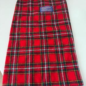 May include: A red and black plaid drawstring bag with a blue label that reads "Pendleton". The bag has a black drawstring closure at the top. The plaid pattern includes green, yellow, and white lines.