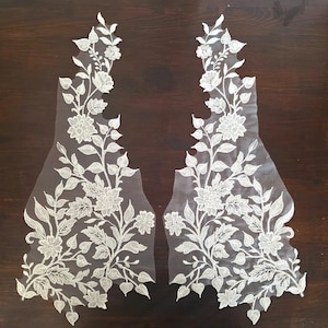 Peut inclure: Deux appliques en dentelle florale blanche avec des motifs brodés complexes de fleurs et de feuilles. Les appliques sont posées sur une surface en bois brun foncé, mettant en valeur les détails délicats de la dentelle.