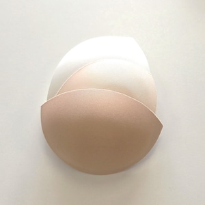 Op de afbeelding: Drie bh-inzetstukken in verschillende nude tinten, van lichtbeige tot een diepere bruintint. De halvemaanvormige inzetstukken zijn ontworpen om in een bh-cup te passen. De achtergrond is een effen wit oppervlak.