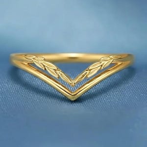 Può includere: Un anello color oro con un design a forma di V. L'anello presenta un delicato motivo a spiga di grano o foglie nella parte superiore della V. L'anello è posto su uno sfondo blu.