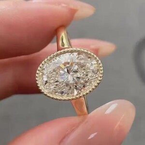 May include: A gold ring featuring a large, oval-cut, clear faceted gemstone set in a bezel with a delicate beaded edge. The band is smooth and tapers toward the setting, showcasing a classic design with a polished finish and a brilliant center stone.